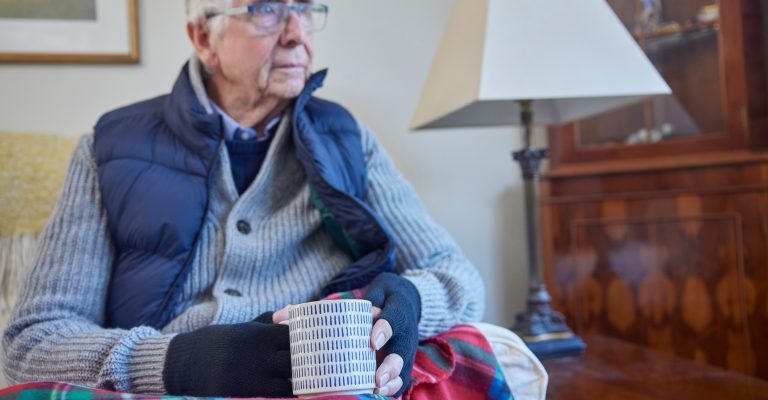 Senior Man Wearing Extra Clothes With Hot Drink Trying To Keep Warm At Home In Energy Crisis